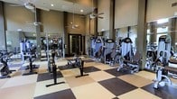 This fitness center is designed to accommodate a comprehensive workout experience within a generously proportioned, single-story layout. The space features soaring ceilings that create an open, airy atmosphere, enhanced by ceiling fans that promote air circulation throughout. The flooring consists of a bold checkered pattern in dark brown and beige, combining durability with a visually striking aesthetic well-suited for high-traffic use. Full-length mirrors line the walls, amplifying the sense of space while allowing users to monitor their form during exercise. The facility is outfitted with a robust selection of strength training equipment, including multiple cable and resistance machines positioned throughout the floor. A rowing machine and weight bench are also present, catering to diverse fitness routines. Recessed lighting supplemented by natural light contributes to a bright and motivating environment. The dark-toned walls provide a focused, gym-like atmosphere that complements the professional-grade equipment. Overall, the space is thoughtfully arranged to maximize usability while maintaining clear pathways between stations, making it functional for simultaneous use by multiple individuals.
