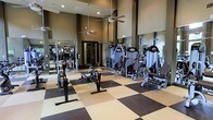 This fitness center is designed to accommodate a comprehensive workout experience within a generously proportioned, single-story layout. The space features soaring ceilings that create an open, airy atmosphere, enhanced by ceiling fans that promote air circulation throughout. The flooring consists of a bold checkered pattern in dark brown and beige, combining durability with a visually striking aesthetic well-suited for high-traffic use. Full-length mirrors line the walls, amplifying the sense of space while allowing users to monitor their form during exercise. The facility is outfitted with a robust selection of strength training equipment, including multiple cable and resistance machines positioned throughout the floor. A rowing machine and weight bench are also present, catering to diverse fitness routines. Recessed lighting supplemented by natural light contributes to a bright and motivating environment. The dark-toned walls provide a focused, gym-like atmosphere that complements the professional-grade equipment. Overall, the space is thoughtfully arranged to maximize usability while maintaining clear pathways between stations, making it functional for simultaneous use by multiple individuals.