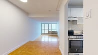 Located on Boulevard De Maisonneuve Ouest in Montreal, this one-bedroom, one-bathroom unit offers a practical and well-organized layout across a single floor. The living area is a bright, open space finished with warm parquet flooring in a classic basket-weave pattern, a hallmark detail carried consistently throughout the unit. Large sliding glass doors open onto a balcony, offering open views of the Montreal skyline and filling the room with natural light. The kitchen is galley-style, featuring white cabinetry reaching to the ceiling, a tiled backsplash, a stainless gas range, and light tile flooring — a clean and functional workspace. A distinctive split-level design separates the bedroom area from the main living space, accessed by a short set of parquet stairs. The bedroom is a well-proportioned private retreat, featuring a window with city views, a closet with a hanging rod, and the same warm parquet flooring. The bathroom is finished with classic white square tile surrounding a full soaking tub and shower combination, complemented by tile flooring. Throughout the unit, white walls and textured ceilings maintain a clean, neutral palette that allows for easy personalization.