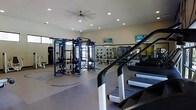 This fitness center is a comprehensively outfitted facility designed to support a full range of workout routines. The space is bright and open, with recessed lighting and a ceiling fan ensuring a comfortable environment during exercise sessions. Large windows invite natural light, contributing to an energizing atmosphere throughout the room. A row of treadmills lines one side of the floor, providing ample cardio options for users. At the center of the space stands a multi-function cable and strength training rig, offering a versatile platform for resistance and functional fitness exercises. A dedicated free weights area, complete with a rack of dumbbells and weight benches, is visible toward the rear of the room. Full-length wall mirrors run along one side, allowing users to monitor their form during workouts. Multiple flat-screen televisions are mounted on the walls, keeping exercisers entertained throughout their sessions. The flooring is a durable, gym-grade surface suited for high-traffic use. The overall layout is thoughtfully organized, ensuring clear zones for cardio, strength training, and free weight exercises, making the facility functional and accessible for a wide variety of fitness needs.