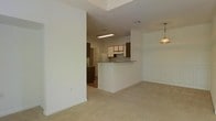 This single-floor space features an open-concept layout that connects the living area to the kitchen, creating a seamless flow between the two spaces. The living area is finished with wall-to-wall carpeting in a neutral beige tone, contributing to a warm and understated ambiance. A pendant light fixture hangs in the corner, adding a subtle decorative touch overhead.

The kitchen is defined by a raised breakfast bar that serves as a natural dividing element between the cooking and living spaces, while still maintaining visual openness. White cabinetry on the lower sections is paired with darker upper cabinets, providing contrast and ample storage. A white refrigerator and additional appliances are visible within the kitchen area, and a flush-mounted fluorescent light fixture illuminates the space from above.

The walls throughout are painted in a crisp white, enhancing brightness and giving the interior a clean, neutral appearance. Architectural details include a recessed ceiling feature above the living area, adding subtle dimension. The overall layout is practical and efficient, making good use of the available space while maintaining an open and airy feel.