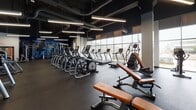 Located at 29 E Green St in Champaign, Illinois, this single-floor fitness center is a well-equipped, modern facility designed to support a comprehensive workout experience. The main cardio and strength area is a standout, featuring an expansive row of Matrix treadmills and elliptical machines positioned along dramatic floor-to-ceiling windows that flood the space with natural light and offer open views of the surrounding neighborhood. Overhead, exposed ductwork and sleek linear pendant lighting against a dark ceiling create an energetic, industrial aesthetic. A dedicated strength training zone features cable machines, squat racks, and a variety of benches, set against bold blue accent walls mounted with flat-screen TVs. A motivational mural with athlete silhouettes and the words "Better, Faster, Stronger" anchors a separate functional training room equipped with cable systems and resistance machines. Additional areas include a flexible studio space with floor-to-ceiling windows, plyo boxes, and mirrored walls, as well as a supplementary room outfitted with a pull-up tower, medicine ball rack, and yoga mats. Throughout, rubber flooring provides safety and durability, while the thoughtful layout ensures distinct zones for cardio, strength, and functional training.