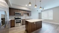 This thoughtfully designed single-floor residence offers a clean, contemporary aesthetic with light wood-style flooring and a neutral color palette carried consistently throughout. The open-concept kitchen serves as the focal point of the living space, showcasing rich dark walnut-toned cabinetry, a decorative arabesque tile backsplash, and a full suite of stainless steel appliances including a range, microwave, dishwasher, and refrigerator. A central kitchen island with a white quartz-style countertop provides additional prep space and casual seating, while stylish pendant lights hang overhead. A ceiling fan near the adjoining living area enhances comfort, and a large window allows natural light to fill the space. A hallway connects the main living area to the private quarters, featuring recessed lighting and panel-style doors. The primary bedroom offers a comfortable footprint with hardwood-style flooring, and direct access to a well-appointed en-suite bathroom. The primary bath includes a dual-sink vanity with dark wood cabinetry, tile flooring, and a glass-enclosed walk-in shower with clean white subway tile. A dedicated walk-in closet with built-in shelving and hanging rods provides ample storage. The second bathroom mirrors the same modern finishes found throughout the residence.