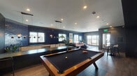 This single-floor amenity lounge is thoughtfully designed to serve as a dynamic social and recreational hub. The space is divided into two distinct yet connected zones: a game room area and a relaxed lounge area, both unified by a cohesive modern aesthetic.

The game room section is anchored by a full-size pool table with dark felt, complemented by a wooden shuffleboard table and a large-format television mounted against a striking black-and-white botanical patterned wallpapered accent wall. Built-in shelving with integrated lighting displays curated décor and artwork, while pendant lighting clusters above the game tables provide focused illumination. A wet bar area with light wood cabinetry and a dark countertop runs along one wall, accented by deep navy glazed brick tiles.

The lounge area transitions seamlessly from the game space, featuring a cognac leather sofa, sculptural navy velvet accent chairs, and a minimalist wood coffee table arranged on a textured area rug. Bold cobalt blue brick-tiled walls serve as a dramatic backdrop, adorned with colorful framed artwork. Natural light filters through multiple windows, and glass doors open to an outdoor terrace, extending the social space beyond the interior. Recessed lighting throughout maintains a warm, inviting ambiance across the entire lounge.