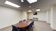 This single-floor conference space is designed to support professional meetings and collaborative work. The room centers around a substantial oval-shaped wood conference table surrounded by blue upholstered chairs, providing comfortable seating for multiple attendees. A wall-mounted flat-screen television serves as the primary display for presentations and video conferencing, complemented by a narrow console table positioned beneath it along the wall.

The space features a drop ceiling with recessed panel lighting and flush-mounted light fixtures that provide consistent, even illumination throughout the room. Patterned loop-pile carpeting in neutral tones covers the floor, adding texture and acoustic dampening to the environment. The walls are finished in a clean off-white tone, contributing to a bright and professional atmosphere.

A wood-framed interior door with a glass panel provides access to the room, while a framed cityscape photograph adds a subtle decorative element to one wall. The layout includes open floor space beyond the table area, offering flexibility for additional furniture or standing room. Overall, the space presents a clean, functional, and well-appointed environment suited for professional use.