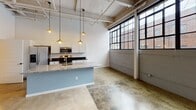 Located at 1255 University Ave in Rochester, NY, this single-floor loft space showcases a striking industrial aesthetic thoughtfully blended with modern finishes. Expansive polished concrete floors extend throughout the open-plan living area, reflecting light and adding to the sense of generous, uninterrupted space. Soaring ceilings with exposed structural beams and visible ductwork reinforce the building's converted industrial character, while large, multi-pane factory-style windows along the exterior wall flood the interior with abundant natural light and offer views of the surrounding brick streetscape.

The kitchen is well-appointed with white shaker cabinetry, dark granite countertops, and a full suite of stainless steel appliances including a side-by-side refrigerator, electric range, and built-in microwave. A granite-topped island with a sink provides additional prep space and casual seating, anchored overhead by a row of warm gold pendant lights that complement the industrial ambiance.

The bathroom is designed with accessibility in mind, featuring wide-format tile flooring, sturdy grab bars, an extended dual-sink vanity with granite countertops and ample cabinetry, and a well-lit mirror. Structural support columns punctuate the open living area, further emphasizing the loft's authentic converted-warehouse character throughout.