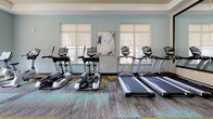 Located at 1209 Westwood Ave in Richmond, Virginia, this single-floor fitness center offers a thoughtfully designed, multi-zone workout facility. The space is divided into distinct areas to accommodate a wide range of exercise needs. A glass-walled spin studio houses several stationary cycling bikes and is outfitted with hardwood-style flooring, a wall-mounted TV, and a large mirror, creating an immersive and energizing environment for cycling classes or individual sessions. The main workout floor features patterned, multi-toned carpet in shades of gray, blue, and yellow, providing a dynamic visual foundation throughout the space. The cardio zone includes multiple treadmills and elliptical machines positioned along a windowed wall, allowing for natural light and outdoor views while exercising. A dedicated strength area offers a full dumbbell rack, flat workout benches, and ceiling-mounted TVs for entertainment. A separate section houses cable and resistance machines suited for targeted strength training. Large windows with roller shades line the perimeter, flooding the space with natural light. Recessed lighting and ceiling fans ensure a comfortable environment throughout. The overall ambiance is modern, clean, and well-organized, reflecting a professional-grade fitness facility.