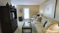 This single-floor space offers a comfortable and thoughtfully arranged living environment. The living room serves as the focal point of the space, featuring a neutral color palette with warm beige walls and plush carpeted flooring that contributes to a relaxed atmosphere. A large, cream-toned sofa anchors the seating area, complemented by decorative zebra-print accent pillows and a matching accent chair with elegant dark wood framing. A glass-top coffee table with a dark wood base sits at the center, adding a refined touch to the room. Opposite the seating arrangement, a substantial dark wood entertainment center houses a television and provides ample storage, with a matching round side cabinet nearby that adds continuity to the design. Natural light flows generously into the room through a glass-paneled door, creating a bright and airy ambiance. The walls are adorned with tasteful artwork, adding character to the space. The overall design aesthetic balances functionality with style, incorporating dark wood furniture accents against light upholstery and neutral walls for a classic, inviting look suitable for everyday living.
