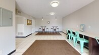 Located in Smyrna, Tennessee, this single-floor community facility at Country Village Drive serves as both a laundry center and a shared common area. The space is thoughtfully divided into two primary zones, unified by clean white tile flooring and neutral gray walls throughout.

The common area greets visitors near the main entrance, where glass-paneled exterior doors allow natural light to fill the space and offer views of the surrounding greenery. Flush-mount ceiling lights provide consistent overhead illumination. The room is furnished with high-top bistro tables and bar stools, as well as a second seating area with teal metal chairs, making it a comfortable gathering spot for residents. A cork bulletin board near the entrance keeps community information readily accessible.

The laundry room is well-equipped with a row of commercial-grade washing machines and dryers, all neatly arranged along the walls and finished in white. Light pours in through a window fitted with blinds, brightening the space naturally. A wall-mounted drying rack provides additional functionality, while tile flooring throughout ensures easy maintenance.

Overall, the facility is clean, practical, and efficiently laid out, offering residents a convenient and welcoming environment for both laundry needs and casual social interaction.