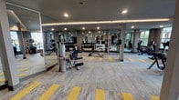 Located at 8233–8241 Meadow Rd in Dallas, TX, this single-level fitness center offers a comprehensive workout environment designed to accommodate a wide range of exercise needs. The space is divided into distinct functional zones, seamlessly connected by a patterned carpet featuring a neutral grey base with bold yellow accent strips that add visual energy to the floor plan.

The strength training area is generously proportioned, housing a variety of weight machines, including cable and resistance equipment from brands such as Hoist, as well as a full dumbbell rack. Floor-to-ceiling mirrors line the walls throughout this zone, amplifying the sense of space and allowing users to monitor their form during exercise. Recessed lighting and ceiling-mounted fans contribute to a comfortable, well-lit environment.

The cardio zone is positioned along a wall of large, framed windows that overlook the surrounding exterior, providing natural light and open views during workouts. Multiple treadmills, elliptical trainers, and stair climbers are arranged in rows facing the windows, creating an airy and motivating atmosphere. Ceiling fans are installed above this area to enhance airflow.

Additional amenities visible within the space include a dedicated area with wood-paneled cabinetry and a display screen, likely serving as an information or check-in station. Overall, the fitness center presents a modern, well-organized, and fully equipped facility.
