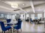 Located at 600 Starkey Rd in Largo, Florida, this 480-square-foot fitness center is a single-floor amenity space designed to support a well-rounded workout routine. The entrance features motivational typography wall graphics displaying words such as "fitness," "agility," "inspiration," and "challenge," setting an energizing tone upon arrival. The main workout area is outfitted with multiple treadmills positioned along a wall of windows that allow natural light to fill the space and offer views of the surrounding greenery. A full cable strength training machine, an adjustable workout bench, a recumbent bike, and an elliptical trainer provide a variety of resistance and cardio options. A dedicated free weights section includes a tiered dumbbell rack and a medicine ball rack stocked with colorful weighted balls. Floor-to-ceiling mirrors line one wall, visually expanding the space and supporting proper form during exercise. A blue yoga mat on the floor designates a stretching and flexibility zone. The flooring throughout consists of durable, multi-toned carpet tiles suited for high-activity use. A textured ceiling with exposed beams adds architectural character, while recessed and flush-mount lighting ensures the space is well-lit at all times. Overall, the fitness center is thoughtfully arranged to maximize functionality within its compact footprint.