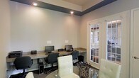 This single-floor space serves as a dedicated business center designed to support productivity and focused work. The room features a built-in countertop workstation that runs along the wall, accommodating multiple desktop computer setups complete with monitors, keyboards, and tower units. Ergonomic office chairs are positioned at each workstation, providing comfortable seating for extended use. The space is finished with neutral, light gray walls that create a clean and professional atmosphere. A distinctive dark-painted tray ceiling border adds a refined architectural contrast, drawing the eye upward and giving the room a sense of elevated design. Recessed lighting is installed throughout the ceiling, ensuring bright, even illumination across the workspace. A glass-topped round table with white upholstered seating is positioned in the foreground, offering an additional area for meetings or casual collaboration. A classic French door with multi-pane glass panels provides access to an adjacent room, allowing natural light to filter in while maintaining a sense of openness. Overall, the space balances functional design with polished finishes, making it well-suited for professional use.