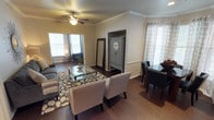 This single-floor space offers a thoughtfully designed open layout that seamlessly connects the living and dining areas, creating a warm and inviting atmosphere. Rich hardwood flooring flows throughout, complemented by crisp white crown molding that adds a classic, refined touch to every room.

The living area is centered around a glass-top coffee table atop a patterned area rug, flanked by plush upholstered seating in neutral tones. A ceiling fan overhead provides both comfort and a subtle decorative element. Large windows dressed with blinds and sheer patterned curtains allow generous natural light to fill the space, enhancing its open, airy feel.

A built-in nook with a sliding barn-style door adds a distinctive architectural detail, offering both character and functional storage. The dining area is appointed with dark wood chairs featuring nailhead trim, situated near additional windows that continue to bring in natural light.

The overall ambiance strikes a balance between casual comfort and tasteful elegance, with neutral wall tones, carefully chosen furnishings, and architectural accents working together to create a cohesive and welcoming environment throughout this single-story home.