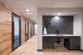 This contemporary single-floor office suite is designed with a refined, modern aesthetic that balances functionality and style. The space is unified by light-toned wood plank flooring in the main corridors and common areas, contrasting elegantly with dark-toned carpet in the conference and private work areas.

The entry corridor serves as a stylish transition zone, featuring a warm wood-clad accent wall that frames a sleek glass-paneled door with black trim. Colorful abstract artwork adorns the walls, lending the space a creative and welcoming atmosphere.

A compact but well-appointed conference room accommodates a small group around a light wood table with tan leather chairs. A wall-mounted flat-screen display makes it ideal for presentations, while a vibrant piece of circular artwork adds visual interest.

A private phone booth or focus pod offers a dedicated space for individual work or confidential calls. Enclosed by a full-height black-framed glass door, the pod features a built-in desk, a single upholstered chair, and a textured statement wall treatment that enhances acoustic privacy and visual depth.

The kitchenette area provides practical amenities including a countertop, sink, and under-counter storage with dark cabinetry, finished with a bold hexagonal tile backsplash. Throughout, recessed lighting and clean white walls contribute to a bright, professional environment.