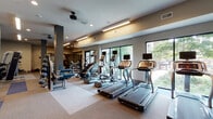 This modern fitness center is thoughtfully designed to support a comprehensive workout experience across its single-floor layout. The main cardio area is lined with large floor-to-ceiling windows that flood the space with natural light and offer views of lush greenery outside, creating an energizing atmosphere. Multiple treadmills, stair climbers, and elliptical machines — each equipped with individual digital display screens — are arranged in organized rows along the windowed wall, making workouts both functional and visually engaging. Ceiling fans and recessed linear lighting complement the bright, open feel of this zone. Adjacent to the cardio section, a dedicated strength training area features a selection of cable machines, weight benches, free weights, and resistance equipment, all set against large wall-mounted mirrors that enhance the sense of space. The flooring transitions between carpet tile and rubberized surfaces, clearly delineating workout zones. A separate studio room provides a more intimate environment, distinguished by a textured geometric accent wall, wall-mounted television, spin bikes, stability balls, resistance bands, and medicine balls. A striking slatted wood architectural feature adds warmth and visual interest to this area. Together, the spaces offer a well-rounded, amenity-rich fitness facility.