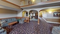 The Residents Lounge is a spacious and warmly appointed communal gathering space designed to foster comfort and community. The room immediately impresses with its soaring vaulted ceiling, which opens the space dramatically and is complemented by an upper mezzanine level with a classic wood-railed balcony overlooking the main floor below. Natural light filters in through transom windows positioned high along the upper walls, brightening the interior alongside warm ambient lighting from table lamps and a chandelier.

A centerpiece tiled fireplace with a wood mantel anchors one side of the main area, adding a cozy, residential charm. The flooring throughout is covered in a rich, patterned carpet in deep jewel tones that ties the space together. Warm wood wainscoting lines the lower portions of the walls, lending a traditional and inviting aesthetic.

Elegant arched passageways divide the lounge into distinct seating alcoves, each furnished with comfortable sofas, armchairs, and end tables — ideal for conversation and relaxation. A carpeted staircase with a turned wood newel post and white balusters provides access to the upper level. Decorative touches such as framed artwork, potted plants, and accent pieces complete the warm, homey atmosphere of this well-designed communal lounge.