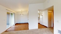 This single-floor residence is thoughtfully designed with a practical, open-concept layout that connects the main living areas seamlessly. The living room features warm wood-look flooring and transitions naturally into a dining area, which is highlighted by a pendant light fixture and a baseboard heating unit along the wall. Large windows and a sliding glass door flood the living space with natural light while providing direct access to an outdoor area with exterior views of surrounding greenery.

The kitchen is positioned adjacent to the dining area and is separated by a partial wall with a pass-through counter, creating an efficient and semi-open workspace. Light wood cabinetry offers generous storage, complemented by white appliances including a refrigerator and electric range, a stainless steel sink, and ample counter space.

The bedrooms are carpeted for comfort and feature neutral walls, standard closets with sliding doors, and windows that allow natural light and outdoor views. Each room offers a quiet, functional environment suitable for a variety of uses.

The bathroom includes a full-size soaking tub with an overhead shower, a curtain rod, and clean white fixtures throughout, maintaining a straightforward and functional design. Overall, the space presents a clean, neutral palette with practical finishes throughout.
