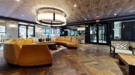 This thoughtfully designed single-floor amenity space makes a strong first impression from the moment you step through its reflective, dark-paneled entry. A stone-tiled foyer transitions seamlessly into the main lounge, where rich herringbone hardwood flooring and a textured concrete ceiling set a sophisticated, contemporary tone throughout.

The central focal point is a sweeping circular sectional sofa in warm amber leather, anchored by a cowhide rug and crowned by a dramatic drum chandelier. This inviting gathering area opens toward large windows that draw in abundant natural light and offer views of the surrounding exterior.

Adjacent to this lounge is a spacious bar and kitchen area, featuring a large dark stone island lined with wooden bar stools and illuminated by a striking row of brass pendant lights. A herringbone wood floor continues here, complemented by a textured concrete ceiling and a large-scale mural that adds artistic character to the space.

A separate seating lounge showcases coffered ceilings with exposed natural wood beams, bold geometric patterned carpet in black, white, and grey, and a curated mix of leather armchairs and settees arranged to encourage conversation. Wall-mounted televisions and decorative sconce lighting enhance the relaxed atmosphere.

A dedicated game area features a marble chess table, upholstered seating, wall sconces, and high-gloss dark wall paneling, rounding out this versatile and richly appointed communal space.
