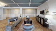 Located at 205 McCaskill Rd E in Pinehurst, NC, this elegantly appointed single-floor space delivers a refined blend of contemporary design and functional versatility. The standout architectural feature is the striking coffered ceiling finished in a deep slate blue, offset by crisp white trim that adds depth and visual interest throughout the open layout. Underfoot, intricate geometric-patterned hardwood flooring in warm natural tones creates a cohesive and polished aesthetic across every zone of the space.

The main lounge area is thoughtfully furnished with a gray sectional sofa, upholstered accent chairs, a bold zebra-print ottoman, and a dark console unit with mirrored accents supporting a wall-mounted television — ideal for relaxed gatherings or community events. A dedicated meeting or co-working zone features a farmhouse-style table with bar seating positioned beneath large windows that invite abundant natural light. Dual sliding barn doors in a dark charcoal finish open to a private conference or office room, adding an industrial-chic element to the overall design.

The space also provides access to a fitness area, visible through glass partitions, and features multiple entry points including a bold black exterior door. Tasteful artwork, lush greenery, and layered area rugs complete the sophisticated, welcoming ambiance throughout.