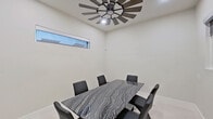 This single-room space presents a clean, contemporary design defined by crisp white walls, large-format porcelain tile flooring, and thoughtful architectural details. The centerpiece of the room is a bold, rectangular table with a dramatic grey-and-white swirling marble surface, surrounded by six dark leather upholstered chairs that reinforce the space's refined, modern aesthetic.

Overhead, a striking windmill-style ceiling fan with wide wooden blades serves as both a functional and decorative focal point, adding a rustic-industrial contrast to the otherwise minimalist interior. Recessed lighting complements the fan's integrated light fixture to ensure the room is well-illuminated.

A horizontal clerestory window along one wall allows natural light to filter in while maintaining privacy, contributing to the room's bright and airy atmosphere. On the opposing wall, a wall-mounted flat-screen television makes the space well-suited for presentations or media viewing. An additional interior pass-through window offers a visual connection to an adjacent space, enhancing the sense of openness.

A solid white paneled door with matte black hardware provides a stylish entry point, consistent with the room's modern design language. The combination of polished surfaces, neutral tones, and carefully chosen fixtures creates a professional yet inviting environment suitable for meetings, dining, or collaborative gatherings.