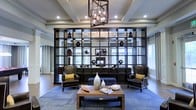 This thoughtfully designed single-floor space makes a strong architectural impression from the moment you enter. The main living area is anchored by a striking, floor-to-ceiling open-grid shelving unit in dark wood, which serves as both a functional display feature and a dramatic room divider, allowing light to pass through while adding depth and visual interest to the space. Curated decorative objects are arranged throughout the shelving, lending the area a gallery-like quality. A geometric chandelier hangs overhead, its warm, exposed bulbs complementing the recessed lighting set within a coffered ceiling that adds a sense of height and structural elegance. The seating arrangement below features a pair of rich leather wingback chairs flanking a central ottoman and a solid wood coffee table, all set atop a softly patterned area rug in cool blue tones. Natural light filters in through large windows dressed with floor-length drapes, enhancing the open, airy feel of the room. The overall ambiance is one of refined sophistication, blending contemporary design elements with classic furnishings to create a space that is both visually compelling and inviting.