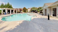 Located at 1600 Jasmine Cove Cir in Simpsonville, South Carolina, this outdoor amenity area offers a well-appointed community pool and spa experience. The centerpiece is a generously sized freeform swimming pool with clear, blue-green water, complemented by an adjacent spa for relaxation. The expansive concrete pool deck provides plenty of open space for sunbathing and recreation, furnished with rows of cushioned lounge chairs arranged for comfortable outdoor leisure.

A white pergola structure anchors one side of the deck, offering a shaded gathering spot accented with colorful patio umbrellas and additional seating. Mature trees and well-maintained landscaping surround the perimeter, creating a lush, natural backdrop and providing natural shade throughout the day. The entire pool area is enclosed by a classic black iron fence with a gated entry, ensuring privacy and security for residents.

Ornamental lamp posts and manicured shrub beds contribute to the polished, welcoming atmosphere of the outdoor space. The surroundings include residential buildings visible in the background, set against open skies that allow abundant natural light to flood the area. This thoughtfully designed outdoor amenity space balances functionality and aesthetics, offering an inviting retreat ideal for warm South Carolina days.