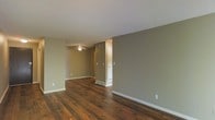 This single-floor space offers a clean, functional layout designed for comfortable everyday living. The interior is finished with rich, dark hardwood floors that run throughout, adding warmth and character to each area. The walls are painted in a soft, neutral taupe tone that complements the flooring and contributes to a calm, cohesive atmosphere.

The main living area is open and spacious, flowing naturally into an adjoining room, creating a connected feel throughout the space. A ceiling fan is installed in the secondary area, providing both practical air circulation and a subtle visual anchor to the room. White baseboards provide a crisp contrast against the taupe walls, adding a polished, finished look to the interior.

A textured popcorn ceiling adds a classic architectural detail overhead, while a doorway with a white-framed door leads to additional areas of the space, offering separation and privacy where needed. Visible floor vents indicate a central HVAC system integrated discreetly into the layout. The overall ambiance is understated and move-in ready, with a neutral palette that serves as an ideal backdrop for a variety of furnishing styles and personal décor preferences.