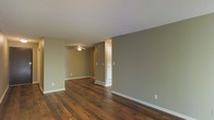 This single-floor space offers a clean, functional layout designed for comfortable everyday living. The interior is finished with rich, dark hardwood floors that run throughout, adding warmth and character to each area. The walls are painted in a soft, neutral taupe tone that complements the flooring and contributes to a calm, cohesive atmosphere.

The main living area is open and spacious, flowing naturally into an adjoining room, creating a connected feel throughout the space. A ceiling fan is installed in the secondary area, providing both practical air circulation and a subtle visual anchor to the room. White baseboards provide a crisp contrast against the taupe walls, adding a polished, finished look to the interior.

A textured popcorn ceiling adds a classic architectural detail overhead, while a doorway with a white-framed door leads to additional areas of the space, offering separation and privacy where needed. Visible floor vents indicate a central HVAC system integrated discreetly into the layout. The overall ambiance is understated and move-in ready, with a neutral palette that serves as an ideal backdrop for a variety of furnishing styles and personal décor preferences.