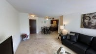 This single-floor residence offers a well-connected open-concept layout that seamlessly integrates the living, dining, and kitchen areas into one cohesive and functional space. The living area is carpeted, providing a warm and comfortable feel underfoot, and is furnished with a leather sofa and accent side table, making it an inviting spot for relaxation. A decorative floral arrangement near the entryway adds a welcoming touch upon arrival through the solid front door. The dining area transitions naturally from the living space, featuring a dark wood dining table and chairs set atop tile flooring, which continues into the kitchen. The kitchen is equipped with dark cabinetry, a sink positioned along the counter, and overhead lighting, offering a practical and efficient cooking environment. A ceiling fan centered above the dining and kitchen area provides both light and air circulation throughout the open space. The consistent neutral color palette of white and grey walls, combined with light-toned ceilings, gives the interior a clean and airy atmosphere. The overall layout is straightforward and efficient, making excellent use of the available space while maintaining a comfortable and livable environment throughout.