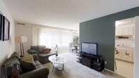 This 1-bedroom, 1-bathroom residence is thoughtfully designed to maximize comfort and functionality within a single-floor layout. The living area serves as the heart of the home, featuring plush wall-to-wall carpeting that adds warmth underfoot. A striking dark teal accent wall creates a bold focal point, providing visual contrast against the neutral tones of the surrounding walls and ceiling. Large windows dressed with vertical blinds span the width of the room, allowing generous natural light to fill the space throughout the day. The open-concept layout seamlessly connects the living area to a dedicated dining space, which is illuminated by a pendant light fixture — ideal for both casual meals and entertaining. The living room offers ample space for full-sized furnishings, demonstrating strong versatility for a variety of arrangements. The overall ambiance strikes a balance between modern style and everyday practicality, with clean lines and a neutral palette that lends itself well to personalization. The thoughtful design and natural light throughout make this a welcoming and functional home.