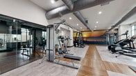 Located at 435 Main St in Hackensack, NJ, this expansive fitness center is a thoughtfully designed, multi-zone facility offering a full suite of workout options for all fitness levels. The space features durable wood-toned and light concrete-style flooring throughout, exposed industrial ductwork along the high ceilings, and recessed lighting that creates a bright, energizing atmosphere.

The main cardio zone is lined with large windows that allow ample natural light and street views, housing a row of treadmills, ellipticals, stair climbers, and rowing machines—all equipped with digital screens. Adjacent to this area, a dedicated strength training zone features a cable machine, weight rack, squat rack, and a multi-station functional trainer, complemented by full-length mirrors that enhance the sense of space.

A separate functional training room with deep teal accent walls hosts spin bikes, medicine balls, kettlebells, resistance bands, and a variety of accessories on organized racks—ideal for group or high-intensity interval training. A striking floor-to-ceiling sunset mural anchors one wall of the main gym area, adding a vibrant, motivating visual element. An additional zone features a scenic bridge mural, a functional trainer, and a stair climber beside large windows. Together, these zones form a comprehensive, well-equipped fitness destination.