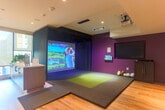 Located at 2230 Calle de Luna in Santa Clara, CA, this single-floor amenity lounge offers a thoughtfully designed recreational space that seamlessly blends entertainment and relaxation. The open floor plan is anchored by warm hardwood flooring throughout, recessed lighting, and a clean white ceiling that lends the space a bright, contemporary feel.

The living area is generously proportioned, featuring a large sectional sofa, marble-topped coffee tables, and a bold textured accent wall in deep purple and grey tones that frames a large wall-mounted television. Floor-to-ceiling windows along multiple walls flood the space with natural light and offer views of the surrounding urban courtyard and neighboring buildings, creating an airy, connected atmosphere.

A standout feature is the fully equipped indoor golf simulator, set within a dedicated dark-framed bay with a projection screen, turf mat, and professional lighting — ideal for recreation and entertainment. Adjacent to the simulator is a casual seating nook with a high-top counter and plush lounge chairs.

Near the entry, a welcoming foyer with additional lounge seating, a vibrant accent wall, potted greenery, and curated artwork establishes an inviting transition into the space. The overall design balances modern aesthetics with functional amenities, making it a versatile and engaging communal destination.