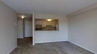 This single-floor residence offers a functional and practical layout designed for comfortable everyday living. The living area is finished with wall-to-wall carpeting, providing a warm and quiet underfoot experience, and is bathed in a neutral color palette throughout, creating a clean and versatile backdrop for any furnishing style.

A defining architectural feature of the space is the pass-through kitchen, which opens directly into the living area via a raised countertop bar. This design promotes an open, connected feel between the cooking and living spaces while maintaining a clear visual boundary. The kitchen is equipped with white cabinetry offering ample storage, a built-in range, a refrigerator, and a sink with countertop workspace along the perimeter. Overhead lighting within the kitchen nook ensures a well-lit cooking environment.

A hallway extending from the living area leads to additional rooms, including what appears to be a private entryway with a closet, providing convenient storage upon arrival. The overall layout is efficient and accessible, with smooth transitions between areas. White walls and neutral tones throughout contribute to a bright, airy ambiance, making the space feel open and easy to personalize.