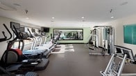 This fitness center offers a comprehensive workout environment designed to support a wide range of exercise routines. The space is finished with durable rubber flooring throughout, providing both safety and comfort underfoot. Recessed lighting illuminates the room evenly, complemented by natural light streaming in through large windows that offer views of the surrounding outdoor landscape.

Along one side of the gym, a row of cardio machines — including treadmills, ellipticals, and stair climbers — is neatly arranged, each equipped with individual display screens. Mounted televisions are positioned for convenient viewing during cardio sessions. A large floor-to-ceiling mirror spans a significant portion of one wall, visually expanding the space and allowing users to monitor their form.

The strength training area features multiple cable and resistance machines, as well as a dedicated free weights section stocked with a full rack of dumbbells, kettlebells, medicine balls, and stability balls. A Rogue-branded bench is also present, catering to more serious lifters. The walls are accented with a soft sage green, lending a calm, motivating ambiance to the room. The overall layout is thoughtfully organized, ensuring ample space to move between equipment zones comfortably.