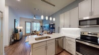 This single-floor residence offers a thoughtfully designed open-concept layout that seamlessly connects the kitchen and living areas. The kitchen is a standout feature, showcasing granite countertops, a large peninsula with bar seating, grey shaker-style cabinetry extending to the ceiling, a herringbone tile backsplash, and a full suite of stainless steel appliances including a refrigerator, range, microwave, and dishwasher. Pendant lighting suspended from a ceiling-mounted track adds a modern touch above the peninsula. Wood-look flooring runs throughout the main living areas, lending warmth and continuity to the space. The open living area is bathed in natural light through large windows and a glass door, complemented by a ceiling fan overhead. Accent walls in a muted blue-grey tone add depth and character to both the living and bedroom areas. The bedroom provides a serene retreat, framed by a recessed accent wall and ample space for a substantial bed. The bathroom features tile-surround tub and shower combination, a granite-topped vanity, wood-look flooring, and direct access to a spacious walk-in closet with built-in shelving. Overall, the residence delivers a clean, contemporary aesthetic with quality finishes throughout.