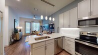 This single-floor residence offers a thoughtfully designed open-concept layout that seamlessly connects the kitchen and living areas. The kitchen is a standout feature, showcasing granite countertops, a large peninsula with bar seating, grey shaker-style cabinetry extending to the ceiling, a herringbone tile backsplash, and a full suite of stainless steel appliances including a refrigerator, range, microwave, and dishwasher. Pendant lighting suspended from a ceiling-mounted track adds a modern touch above the peninsula. Wood-look flooring runs throughout the main living areas, lending warmth and continuity to the space. The open living area is bathed in natural light through large windows and a glass door, complemented by a ceiling fan overhead. Accent walls in a muted blue-grey tone add depth and character to both the living and bedroom areas. The bedroom provides a serene retreat, framed by a recessed accent wall and ample space for a substantial bed. The bathroom features tile-surround tub and shower combination, a granite-topped vanity, wood-look flooring, and direct access to a spacious walk-in closet with built-in shelving. Overall, the residence delivers a clean, contemporary aesthetic with quality finishes throughout.