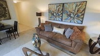 This single-floor living space centers around a spacious, well-appointed living room designed with comfort and style in mind. The room is anchored by a generously sized brown suede sofa adorned with an assortment of decorative pillows in neutral tones of gray, white, and copper, creating a warm and inviting atmosphere. A pair of large botanical-themed framed artworks hang prominently above the sofa, adding a bold visual focal point with earthy tones of blue, gold, and brown. The living area features soft, neutral-toned carpeting that extends throughout, contributing to the overall warmth of the space. A sculptural glass coffee table accessory and a uniquely designed table base add artistic character to the room. Ambient lighting is provided by a sleek floor lamp with a dark shade, casting a soft, warm glow. The walls are finished in a smooth, light neutral tone that complements the warm furnishings. The overall ambiance of the space is relaxed yet refined, offering a comfortable environment with thoughtfully curated décor elements that tie the room together cohesively.