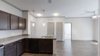 This single-floor residence offers a thoughtfully designed open-concept layout that seamlessly connects the kitchen and living areas, creating a spacious and functional environment. The kitchen is a standout feature, showcasing rich dark wood cabinetry that extends to the upper walls, complemented by granite countertops with subtle veining. A peninsula with an integrated stainless steel sink serves as both a functional workspace and a natural dividing point between the kitchen and the adjoining living space. Pendant lighting hangs above the peninsula, adding a modern, polished touch to the area. The kitchen is also equipped with a built-in dishwasher, maximizing convenience. Durable wood-look flooring runs throughout the open areas, providing a warm and cohesive aesthetic. The neutral white walls and light-toned ceilings enhance the brightness of the space, while multiple HVAC vents ensure year-round comfort. Gray interior doors with raised paneling add a clean, contemporary design element throughout. The open sight lines from the kitchen into the living area and beyond create an airy, connected feel, making the space ideal for both everyday living and entertaining.