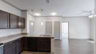 This single-floor residence offers a thoughtfully designed open-concept layout that seamlessly connects the kitchen and living areas, creating a spacious and functional environment. The kitchen is a standout feature, showcasing rich dark wood cabinetry that extends to the upper walls, complemented by granite countertops with subtle veining. A peninsula with an integrated stainless steel sink serves as both a functional workspace and a natural dividing point between the kitchen and the adjoining living space. Pendant lighting hangs above the peninsula, adding a modern, polished touch to the area. The kitchen is also equipped with a built-in dishwasher, maximizing convenience. Durable wood-look flooring runs throughout the open areas, providing a warm and cohesive aesthetic. The neutral white walls and light-toned ceilings enhance the brightness of the space, while multiple HVAC vents ensure year-round comfort. Gray interior doors with raised paneling add a clean, contemporary design element throughout. The open sight lines from the kitchen into the living area and beyond create an airy, connected feel, making the space ideal for both everyday living and entertaining.