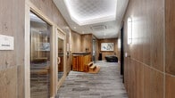 Located at 900 Second Ave N in Nashville, TN, this single-floor wellness suite is a thoughtfully designed recovery and relaxation destination. The space is unified by a cohesive aesthetic of warm wood-paneled walls and large-format stone-look porcelain tile flooring that runs throughout, creating a spa-like atmosphere that is both contemporary and inviting.

The central hallway serves as the spine of the suite, connecting each wellness zone with a sense of purposeful flow. Recessed and cove lighting casts a soft, ambient glow along the corridor, complementing the natural wood tones of the walls. A glass-doored infrared sauna is prominently featured along the hallway, fitted with slatted cedar benches and accessible via wooden step platforms. Adjacent to the sauna, a cedar-clad cold plunge tub with teak steps offers a contrast therapy experience.

The bathroom area extends the design language of the suite, incorporating pebble stone accent walls, open shelving, and clean grey cabinetry topped with a white surface. A dedicated red light therapy room, bathed in warm amber tones, houses full-panel LED therapy devices alongside lush potted plants and a comfortable lounge chair. The suite also includes a reclined massage or relaxation chair positioned beneath a wall-mounted art piece, completing a well-rounded, full-body wellness environment.