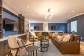 Located at 7820 Bolsa Ave in Midway City, California, this single-floor space presents a sophisticated lounge environment designed with a coastal California aesthetic. The open layout is anchored by light hardwood flooring that flows throughout, complemented by crown molding and recessed lighting paired with an elegant multi-arm chandelier that serves as a central focal point.

The main lounge area features a rich brown leather sofa and mixed-material seating arranged around a dark oval coffee table, all set atop a textured area rug. A full built-in entertainment wall with dark cabinetry, open shelving, and a mounted flat-screen TV spans one side of the room, while a bold navy blue accent wall frames a second TV and a dedicated seating and workspace area with small dining tables and upholstered chairs.

A secondary nook provides a more intimate setting, furnished with a round bistro-style table and chairs beneath a pair of framed coastal photography prints. Dark open shelving with decorative accessories adds visual depth to this area. A third zone features a curved navy wall with large windows, allowing natural light to filter in while maintaining a sense of privacy. The overall ambiance is polished, welcoming, and cohesive throughout.