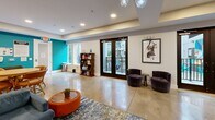 Located at 1325 Boland Pl in Richmond Heights, Missouri, this single-floor amenity space offers a thoughtfully designed open-concept layout combining a fully equipped kitchen and a spacious living area. Polished concrete floors run throughout, lending the space a contemporary, industrial-chic character that complements the modern design aesthetic.

The kitchen is a standout feature, boasting sleek white cabinetry, stainless steel appliances including a large French-door refrigerator, and a dramatic mosaic mirror tile backsplash that adds a striking visual focal point. Pendant lighting hangs above multiple butcher-block communal tables with metal bar stools, creating an inviting atmosphere for gathering and dining.

The living area is anchored by a large wall-mounted flat-screen television with flanking wall-mounted speakers, set against a warm terracotta-toned accent wall adorned with framed artwork. Plush seating, including a generous sectional sofa and swivel lounge chairs, is arranged around a patterned area rug, offering comfortable spaces for relaxation or socializing. A vibrant teal accent wall adds a bold pop of color, while large glass doors open to an outdoor balcony, flooding the space with natural light. Additional seating and game areas round out this well-appointed communal space.