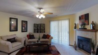 This single-story space centers around a comfortable living room that combines functional design with a relaxed ambiance. The room features a textured ceiling fitted with a ceiling fan and light fixture, providing both air circulation and overhead lighting. A large sliding glass door, dressed with full-length vertical blinds, serves as a prominent architectural element along one wall, allowing natural light to filter into the space while offering privacy control and potential outdoor access. The warm yellow-toned walls add a welcoming feel to the room. Flooring transitions from hard surface near the entryway to plush carpeting throughout the main living area, with a decorative area rug anchoring the seating arrangement. The living room comfortably accommodates a sofa and loveseat, accented with decorative throw pillows, and is complemented by a low-profile coffee table. Wall art and a decorative room divider panel add visual interest, while a small fireplace mantel in the corner provides an additional architectural detail. Overall, the space offers a practical and inviting layout suited for everyday living.