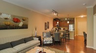 This single-floor space features an open-concept layout that seamlessly connects the living area, dining space, and kitchen into one cohesive environment. The living area is anchored by a comfortable striped sofa and an upholstered accent chair, complemented by a soft area rug and a coffee table, creating an inviting setting for relaxation. Warm earth tones carry throughout, lending a cozy and welcoming atmosphere.

Adjacent to the living area, a round dining table surrounded by cushioned chairs provides an intimate spot for meals. The space transitions naturally into the kitchen, which features warm wood cabinetry, stainless steel appliances including a full-size refrigerator, and a raised breakfast bar with seating. Pendant lighting above the kitchen adds a refined touch, while a track lighting system illuminates the open living and dining areas.

Crown molding lines the ceilings, adding a subtle architectural detail that elevates the overall aesthetic. Rich hardwood-style flooring runs throughout, tying the rooms together cohesively and contributing to the warm, inviting character of the space.