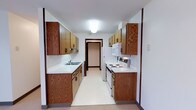 This single-floor space centers around a well-organized galley-style kitchen designed for efficient use of space. The kitchen features warm-toned oak wood cabinetry on both sides, offering generous upper and lower storage throughout. The cabinetry is accented by dark hardware, providing a classic contrast against the white countertops that run along each wall.

On one side, a stainless steel sink is seamlessly integrated into the countertop, accompanied by a textured white tile backsplash that adds subtle visual interest. The opposite wall houses white appliances, including an electric coil stove, an over-the-range microwave, and a full-sized refrigerator, keeping all cooking essentials neatly aligned. The light-colored vinyl flooring reflects the overhead flush-mount ceiling light, keeping the narrow corridor feeling bright and clean.

A doorway at the far end of the kitchen provides a sense of depth and connects to an adjacent area of the home. The overall design is functional and straightforward, with neutral tones and traditional woodwork creating a practical, comfortable kitchen environment suited to everyday use.