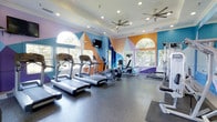 The fitness center at Eagle Rock at Freehold is a well-appointed, single-floor exercise facility designed to inspire and energize its users. The space immediately captures attention with its bold, geometric wall mural rendered in vivid shades of blue, orange, teal, and purple — creating a dynamic and motivating backdrop for workouts. Large arched windows line the walls, flooding the room with abundant natural light and offering pleasant views of the surrounding greenery outside. The tray ceiling adds architectural definition to the space, while dual ceiling fans ensure comfortable air circulation throughout. The fitness area is equipped with a row of treadmills, an elliptical trainer, a recumbent bike, and a cable strength machine, providing a well-rounded selection of cardio and resistance training options. A wall-mounted television is positioned for easy viewing during workouts. The flooring is a durable, dark rubberized surface well-suited for an active environment. The overall ambiance strikes a balance between vibrant energy and functional design, making it an inviting space for residents looking to maintain an active lifestyle.