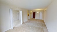 This single-floor residential space offers a clean, functional layout designed to maximize comfort and usability. The main living area is open and spacious, featuring light-toned flooring that extends throughout, contributing to a bright and airy atmosphere. Smooth, neutral-painted walls and a white ceiling create a cohesive, understated aesthetic throughout the unit.

A pendant light fixture hangs near the entryway, providing a subtle decorative accent above the rich, dark-toned front door, which serves as a welcoming focal point upon entry. A large closet flanks the entryway, offering generous storage directly accessible from the main living space.

Adjacent to the living area, the kitchen is partially visible and features warm-toned cabinetry with white appliances, providing a functional cooking space that connects naturally to the rest of the layout. Additional doors visible from the main room suggest access to further spaces, such as bedrooms or utility areas, indicating a multi-room floor plan.

The overall ambiance of the space is calm and neutral, making it well-suited for a variety of furnishing styles and personal tastes. The straightforward layout, ample natural light, and consistent finishes throughout give the space a practical yet comfortable feel that appeals to everyday living.