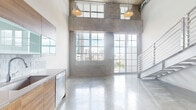 Located at 1657 N Miami Ave in Miami's dynamic Wynwood/Edgewater corridor, this two-story loft showcases a bold industrial aesthetic defined by exposed concrete ceilings, raw concrete block walls, and polished concrete floors throughout. The main living area commands attention with soaring double-height ceilings, oversized grid-pane windows and glass doors that flood the space with natural light and frame striking Miami skyline views. Pendant lighting and a sleek metal staircase with horizontal rail detailing connect the two levels with modern precision. The kitchen is outfitted with warm wood-grain cabinetry, frosted glass upper cabinets, white subway tile backsplash, stainless steel appliances including a French door refrigerator, wall oven, and dishwasher, and a stainless countertop with integrated sink. A bright, open corridor with exposed red pipe accents and a concrete ceiling leads to additional rooms, including a well-proportioned bedroom and a full bathroom featuring a wood-look tile floor, wood-grain vanity, and a fully tiled glass-enclosed walk-in shower. An in-unit stacked washer and dryer adds everyday convenience. The upper level offers open loft space overlooking the main floor, completing this urban retreat.