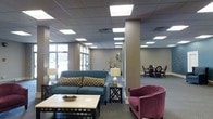 This single-floor space presents an open, well-appointed common area that seamlessly blends a lounge and dining zone into a cohesive, functional environment. The interior features a drop-panel ceiling outfitted with recessed fluorescent lighting panels that provide consistent, bright illumination throughout. Large windows along one wall invite natural light, enhancing the airy atmosphere.

The lounge area is anchored by a teal upholstered sofa adorned with patterned accent pillows, complemented by a plush, studded magenta armchair that adds a bold pop of color. A square coffee table with decorative trim serves as the centerpiece, while a side table with a lamp provides warm supplemental lighting. A structural column with a neutral finish divides the space without disrupting its open feel.

Beyond the lounge, a dining area is furnished with round tables and upholstered chairs in deep purple tones, creating an intimate setting suited for group gatherings. The neutral beige and gray wall palette unifies both areas, while decorative accents—including a sculptural wall mirror and floral arrangement—add refined character. The carpeted flooring ties the space together with warmth and practicality.