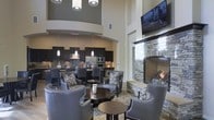 This stylish single-floor space exudes a modern, upscale ambiance with a thoughtfully designed open-concept layout. The dramatic two-story ceilings create an expansive, airy feel, drawing the eye upward to a curved architectural detail that adds a refined architectural touch. A striking stacked-stone fireplace serves as a natural focal point in the lounge area, complemented by a wall-mounted flat-screen television above, making it an ideal setting for relaxed gatherings. The seating area is furnished with elegant grey upholstered armchairs featuring nailhead trim, arranged around dark circular accent tables atop a plush area rug — lending a sophisticated, lounge-like atmosphere. Pendant lighting and recessed fixtures illuminate the space with a warm, inviting glow. The open kitchen at the rear showcases dark espresso cabinetry, stainless steel appliances including a double wall oven and refrigerator, and a stylish mosaic tile backsplash. A raised bar counter with seating provides an additional casual dining option. The neutral color palette of warm beige walls, dark furnishings, and rich stone accents ties the entire space together into a cohesive, contemporary design that is both functional and visually impressive.